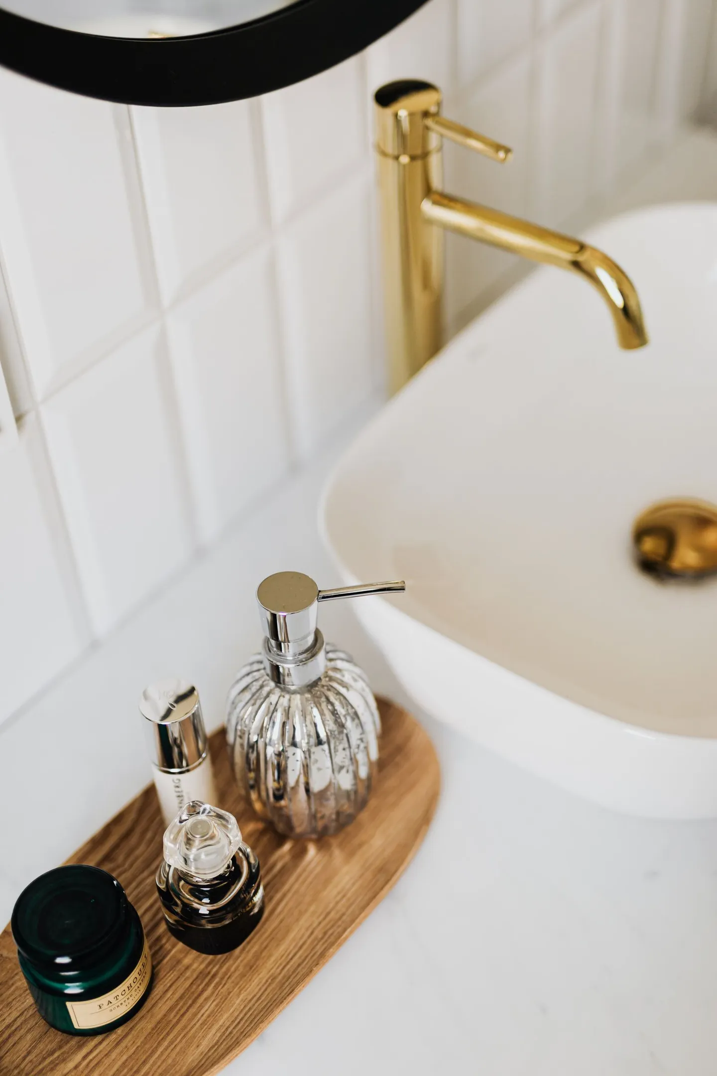 Glass soap dispenser and toiletries on a wooden tray beside a white sink and brass faucet.